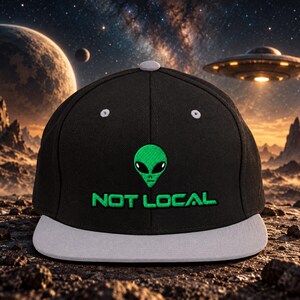 Könnte beinhalten: Eine schwarze Baseballkappe mit hellgrauem Schirm und oberem Knopf. Die Vorderseite zeigt einen gestickten grünen Alien-Kopf über dem Text "NOT LOCAL" in passender grüner Schrift. Die Kappe hat weiße Belüftungsösen an der Krone.