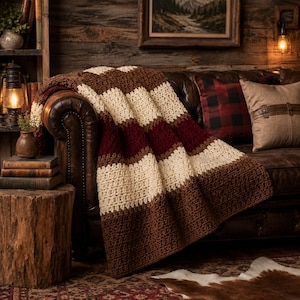 May include: A crocheted throw blanket featuring wide horizontal stripes in alternating colors of dark brown, cream, and deep burgundy. The textile has a textured, repeating stitch pattern throughout, draped over the arm of a dark leather sofa.