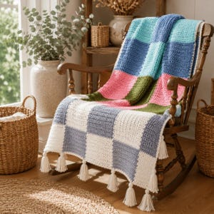 Può includere: Una coperta all'uncinetto con un motivo a scacchi nei toni dell'azzurro, blu scuro, rosa, verde oliva, bianco e grigio. La coperta è drappeggiata su una sedia a dondolo in legno ed è rifinita con nappe color crema lungo il bordo inferiore.