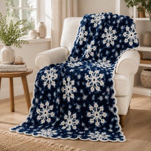May include: A dark blue and white crochet blanket featuring a repeating pattern of large, intricate white snowflakes. The blanket has a scalloped edge and is draped over a cream-colored armchair, showcasing a textured, handmade yarn construction.