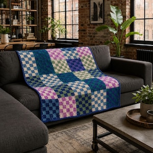 Könnte beinhalten: Eine Häkeldecke mit einem Schachbrettmuster aus quadratischen Blöcken in den Farben Dunkelblau, Petrol, Hellgrün und sanftem Violett. Das Textil zeigt ein durchgehendes Rasterdesign mit abwechselnden Farben über die gesamte Oberfläche.