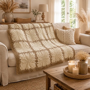 May include: A cream-colored crochet blanket featuring a grid of square motifs joined by a brown yarn border. The blanket is draped over a sofa and displays a textured, repeating floral pattern within each square, finished with a brown crocheted edge.