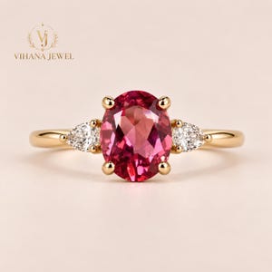 Puede incluir: Un anillo de oro con una gran piedra preciosa rosa de corte ovalado en el centro, sujeta por cuatro garras. A cada lado de la piedra central, hay una piedra preciosa transparente más pequeña de corte en forma de pera. El aro es de oro liso.