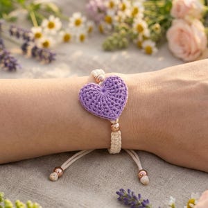 Puede incluir: Una pulsera con un motivo central de corazón tejido a ganchillo en color lavanda. La banda está hecha de cordón color hueso, acentuada con pequeñas cuentas metálicas de oro rosa situadas cerca del corazón y en los extremos del cierre.