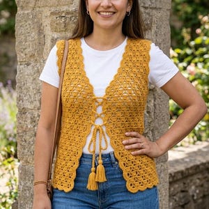 May include: A mustard yellow sleeveless crochet vest featuring an open-front design with a lace-up closure, decorative tassels at the ends of the ties, and a scalloped edge trim, worn over a plain white short-sleeved t-shirt and blue denim jeans.
