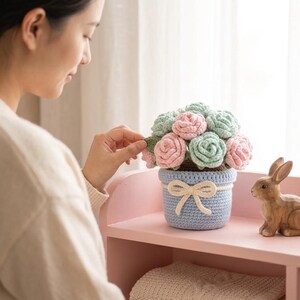 Puede incluir: Un arreglo floral de ganchillo con rosas de color rosa pastel y verde menta en una maceta de ganchillo azul claro. La maceta está decorada con un lazo de hilo color crema atado alrededor del borde. Una pequeña figura de conejo de madera marrón.