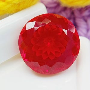 May include: A round, vibrant red gemstone featuring a complex, multi-faceted cut that creates a symmetrical, star-like pattern across its surface. The stone is translucent, allowing light to pass through and highlight its deep crimson colour.