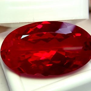 May include: A vibrant, oval-cut red gemstone with a faceted surface that reflects light, displayed resting inside a white plastic jewelry box. The stone exhibits a deep, saturated crimson hue and a polished finish, highlighting its clear, transparent structure.