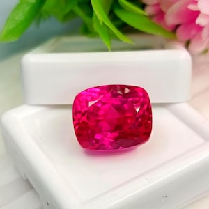 May include: A vibrant, cushion-cut gemstone with a deep, saturated pinkish-red hue. The stone features a faceted surface that reflects light, creating a brilliant appearance. It is displayed resting on a white, square-shaped plastic storage case.