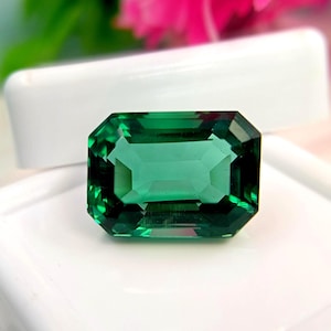 May include: A loose emerald-cut gemstone with a deep, vibrant green color and a faceted surface that reflects light. The rectangular stone is positioned in front of a white display case, showcasing its transparent clarity and sharp, clean edges.