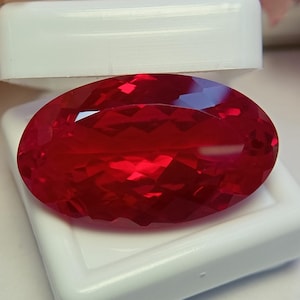 Può includere: Una gemma rossa vibrante dal taglio ovale con una superficie sfaccettata che riflette la luce, esposta all'interno di una piccola scatola rettangolare in plastica bianca con un coperchio incernierato parzialmente aperto.