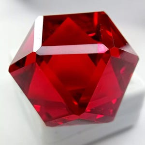 May include: A faceted, vibrant red gemstone with a geometric, multi-sided cut. The stone features sharp edges and polished surfaces that reflect light, creating deep crimson highlights and shadows throughout its transparent, crystalline structure.