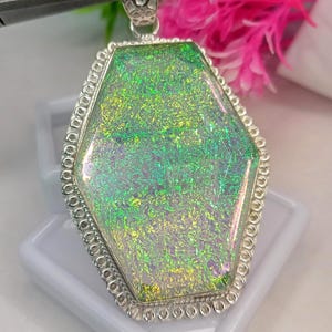 Può includere: Un ciondolo a forma di bara con un inserto in vetro dicroico iridescente dai riflessi verdi e dorati. Il vetro è incastonato in una cornice in metallo color argento con un bordo decorativo a perline e una contromaglia superiore testurizzata.