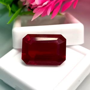 Peut inclure: Une pierre précieuse rectangulaire à facettes d'un rouge profond, dotée d'angles vifs et d'une surface polie. La pierre est placée devant un boîtier de présentation en plastique blanc, avec des éléments floraux rose vif en arrière-plan.