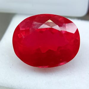 Peut inclure: Une pierre précieuse de taille ovale d'un rouge vif avec une surface à facettes qui reflète la lumière. La pierre présente une forme symétrique avec de multiples facettes triangulaires et en losange sur le dessus, créant un aspect brillant.