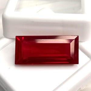 May include: A rectangular, faceted gemstone with a deep, vibrant red colour. The stone features sharp, clean edges and a polished surface that reflects light, showcasing its transparent quality. It is positioned against a white display case.