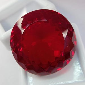 Könnte beinhalten: Ein runder, facettierter Edelstein in einer tiefen, leuchtend roten Farbe. Der Stein weist einen präzisen Schliff mit vielen Facetten auf, der ein symmetrisches Muster aus hellen und dunklen Reflexionen auf der Oberfläche erzeugt.