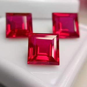May include: Three square-cut, vibrant red gemstones with faceted surfaces. The stones are arranged in a triangular formation, displaying deep crimson hues and sharp, geometric edges that reflect light across their polished, transparent faces.