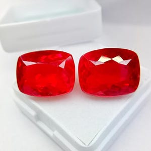 Könnte beinhalten: Zwei leuchtend rote Edelsteine im Kissenschliff mit facettierten Oberflächen, die das Licht reflektieren. Die losen Steine liegen nebeneinander auf dem weißen Schaumstoffeinsatz einer offenen, rechteckigen weißen Kunststoff-Aufbewahrungsbox.