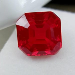 May include: A vibrant, square-cut red gemstone with a faceted surface that reflects light. The stone features sharp, clean edges and a deep, saturated crimson hue, showcasing a symmetrical geometric design with multiple internal light reflections.