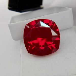 May include: A vibrant, deep red gemstone with a cushion cut, featuring multiple facets that reflect light. The gemstone is positioned in front of a white plastic container with a black lid, showcasing its intense color and geometric clarity.