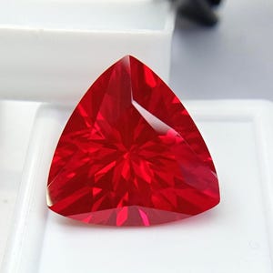 Könnte beinhalten: Ein leuchtend tiefroter Edelstein in dreieckiger Form mit zahlreichen Facetten, die ein sternförmiges Lichtreflexionsmuster erzeugen. Der Stein ist durchscheinend und zeigt eine brillante, gesättigte karmesinrote Farbe auf seiner gesamten Oberfläche.