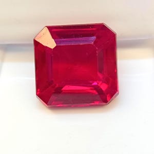 Puede incluir: Una piedra preciosa de color rojo vibrante con corte cuadrado y superficies facetadas que reflejan la luz. La piedra presenta una muesca o inclusión de color claro visible en la esquina superior izquierda, mostrando un tono carmesí saturado.