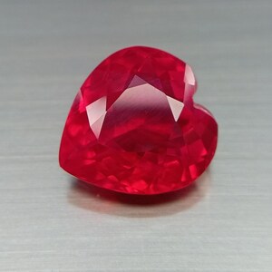 May include: A heart-shaped gemstone with a vibrant, deep red color and a faceted surface. The stone features multiple triangular and diamond-shaped facets that reflect light, creating a brilliant appearance with sharp edges and a pointed base.