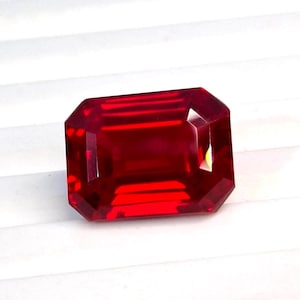 Può includere: Una gemma di colore rosso intenso e vibrante, tagliata in una forma rettangolare a smeraldo. La pietra presenta sfaccettature nette e pulite che riflettono la luce, creando un aspetto brillante e saturo con una chiara trasparenza interna e una superficie levigata.