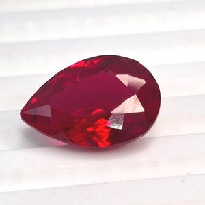 Può includere: Una gemma sfaccettata a forma di pera di un colore rosso intenso e vibrante. La pietra presenta una superficie lucida con molteplici sfaccettature geometriche che riflettono la luce, creando riflessi luminosi e profonde tonalità rosse.