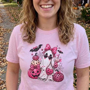 Puede incluir: Una camiseta de manga corta de color rosa claro con un estampado gráfico de un fantasma blanco con un lazo rosa y gafas de sol en forma de corazón, rodeado de calabazas rosas, una bebida rosa, murciélagos y telarañas en tonos rosa y negro.