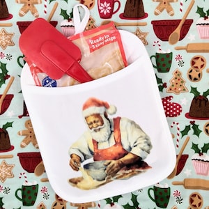 May include: A white fabric oven mitt featuring a printed illustration of Santa Claus wearing an apron and mixing dough in a bowl. The mitt contains a red silicone spatula and a clear plastic pouch with text reading Ready in 3 easy steps.