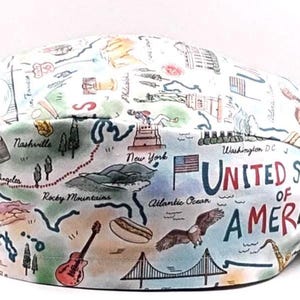 May include: A white and light blue scrub cap featuring a colorful map of the United States. The map includes illustrations of landmarks, a guitar, a hot dog, and an eagle. The words "UNITED STATES OF AMERICA" are prominently displayed.