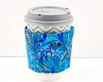 May include: A disposable paper coffee cup with a gray plastic lid and a gray and white chevron pattern on the upper portion. The cup is wrapped in a fabric sleeve featuring a vibrant, swirling marbled design in shades of blue, teal, and purple.