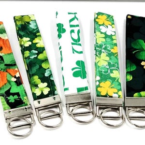 May include: Five fabric wristlet keychains with silver-tone metal hardware and split rings. The designs feature St. Patrick's Day themes including orange and green leprechaun hats, various green shamrock patterns, and one white strap with green text.