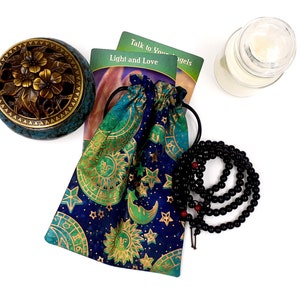 K&ouml;nnte beinhalten: Eine blaue und gr&uuml;ne Kordeltasche mit goldenen Himmelsmotiven, eine schwarze Perlenkette, ein kleiner R&auml;ucherofen und zwei Orakelkarten mit dem Text "Sprich mit deinen Engeln" und "Licht und Liebe".