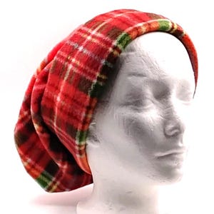 May include: A slouchy beanie hat made of soft, fuzzy fabric featuring a vibrant red, green, and white plaid tartan pattern. The hat is displayed on a white mannequin head, showing a relaxed, rounded shape that covers the crown and back of the head.