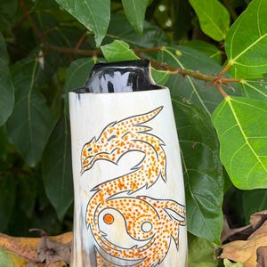 May include: A drinking horn with a polished white and tan surface featuring a black-outlined dragon design filled with orange dots. The dragon curves around a central yin-yang symbol. The top rim is finished in solid black, creating a contrast.