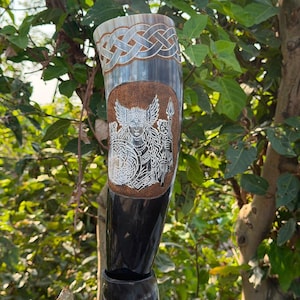 Puede incluir: Un cuerno para beber curvo hecho de material oscuro pulido con un intrincado diseño grabado en blanco de una figura alada que sostiene una lanza y un escudo, rodeado por un fondo de color cobre punteado y un borde de nudos celtas.