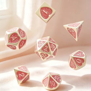 Può includere: Un set di sette dadi poliedrici con bordi in metallo dorato e inserti a forma di cuore riempiti di glitter rosa. Ogni faccia mostra un numero bianco, inclusi un d20, d12, d10, d8, d6, d4 e un dado percentuale.