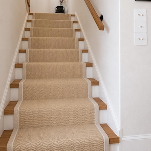 May include: A staircase featuring light brown wooden steps and a central beige runner with a herringbone pattern. The stairs include a wooden handrail on the right side, white walls, and a black vase with floral branches at the top landing.