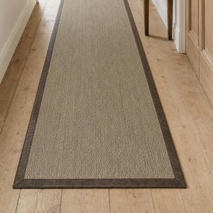 May include: A long, rectangular runner rug featuring a light beige, woven herringbone pattern in the center, framed by a solid, dark brown border. The rug is positioned lengthwise on a light-colored wooden floor, extending into the hallway.