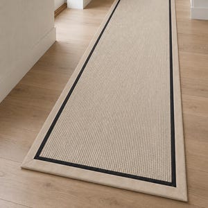 Peut inclure: Un long tapis de couloir rectangulaire doté d'un centre tissé beige clair texturé. Le tapis est encadré par une fine bordure noire distincte, elle-même entourée d'une bordure en tissu beige clair plus large sur les quatre côtés.