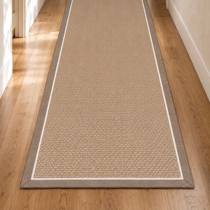 Puede incluir: Una alfombra de pasillo rectangular larga con un centro tejido en color tostado texturizado, un borde interior blanco fino y un borde exterior más ancho en gris sólido. La alfombra está colocada a lo largo sobre un suelo de madera.