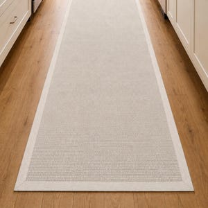 Peut inclure: Un long tapis de couloir rectangulaire doté d'un panneau central beige clair texturé avec un motif tissé subtil, encadré par une bordure unie et lisse blanc cassé. Le tapis est placé dans le sens de la longueur sur un parquet en bois.