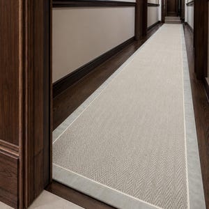 May include: A long, rectangular runner rug featuring a light grey herringbone pattern in the centre, bordered by a thin white line and a wider, solid light grey fabric edge. The rug is positioned lengthwise along a dark wood floor in a hallway.