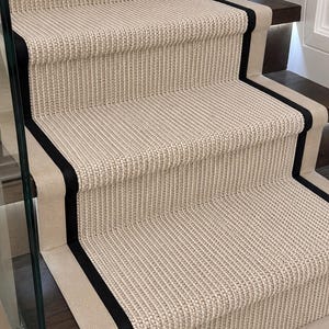 Puede incluir: Una alfombra para escaleras de tejido beige texturizado con un borde negro prominente. La alfombra está instalada sobre peldaños de madera marrón oscuro, con un panel de vidrio visible en el lado izquierdo e iluminación integrada.