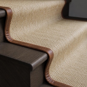 May include: A stair runner featuring a textured beige woven material with a subtle herringbone pattern. The runner is finished with a smooth, dark brown leather-look binding along the edges and is installed on dark wood staircase steps.