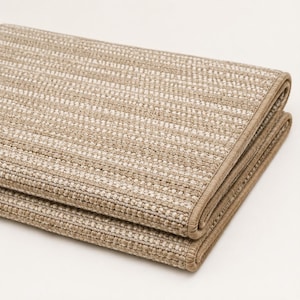 Puede incluir: Una alfombra rectangular plegada que presenta un tejido texturizado con rayas alternas en beige y blanco roto. Los bordes están rematados con un ribete de tela beige a juego, creando un borde limpio alrededor del material tejido.