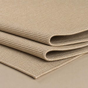 May include: A folded beige area rug featuring a dense, uniform woven texture with a consistent grid-like pattern. The edges of the rug are finished with a matching bound border, showcasing the thickness and structured weave of the material.